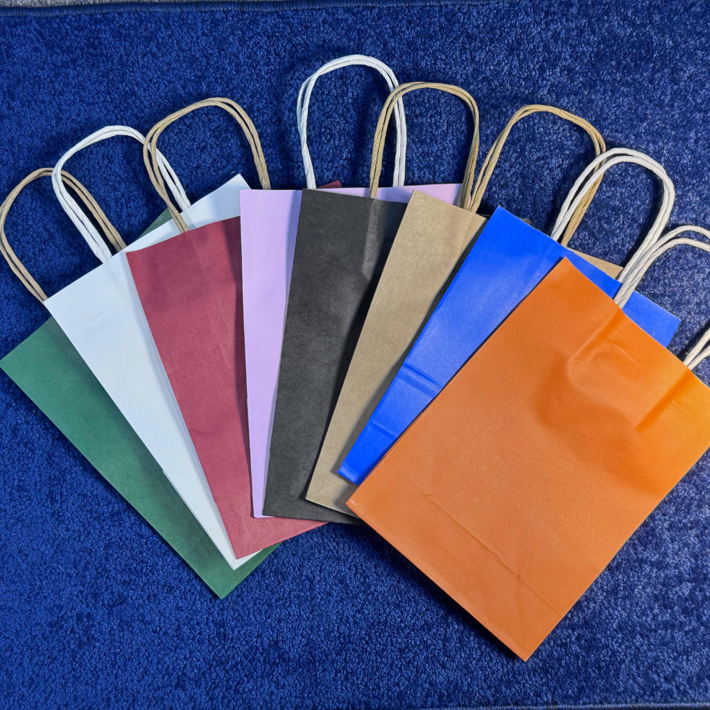 Colorful assortment of A6-sized kraft paper bags with sturdy handles, displayed on a blue textured background. Includes bags in green, white, maroon, black, pink, gold, blue, and orange hues, ideal for versatile packaging needs